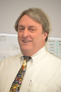 Fair skin main in off-white button shirt and patterned tie smiling and standing in front of a dry erase board