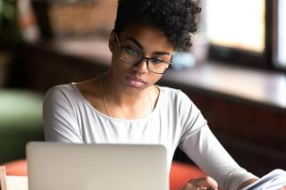 Thoughtful African American student in glasses writing while using laptop computer